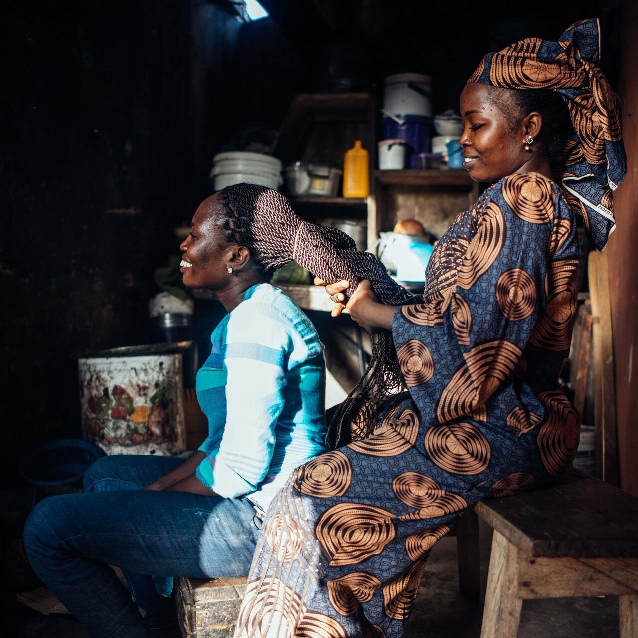 A young woman braids a friend's hair in a Lagos market salon · the everyday ritual at the heart of African beauty